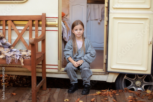 Smiling little girl in casual clothes sitting on porch RV house in garden. Cute young girl sitting near trailer door. Child in cozy campsite fall backyard. Concept camping, outdoor, nature, adventure	