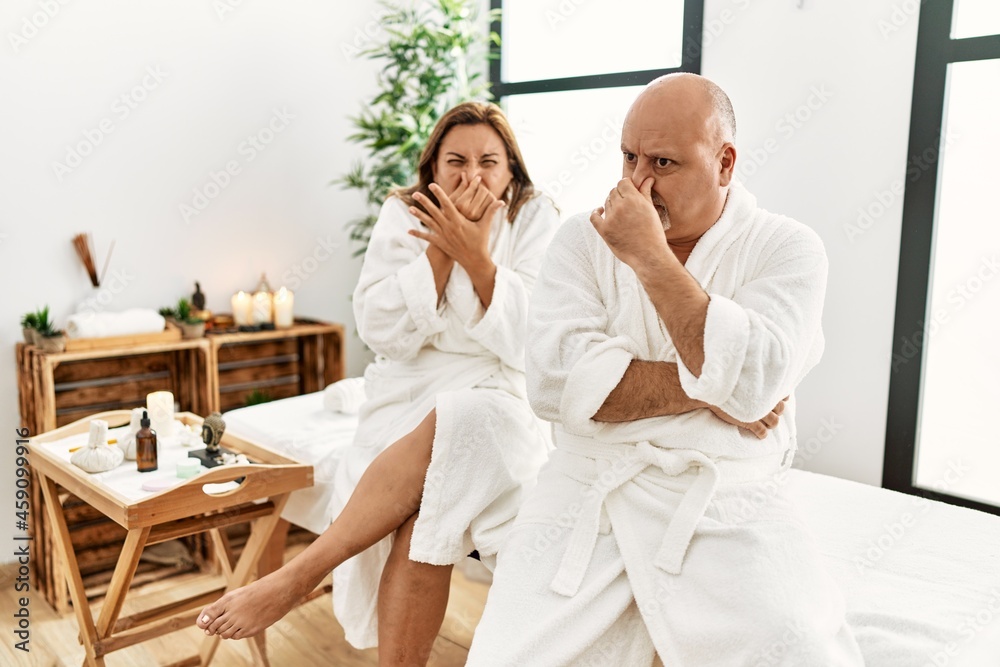Middle age hispanic couple wearing bathrobe at wellness spa smelling something stinky and disgusting, intolerable smell, holding breath with fingers on nose. bad smell Middle age hispanic couple wearing bathrobe at wellness spa smelling something stinky and disgusting, intolerable smell, holding breath with fingers on nose. bad smell