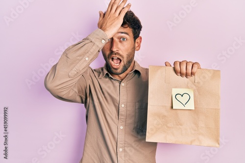 Handsome man with beard holding delivery paper bag with heart reminder surprised with hand on head for mistake, remember error. forgot, bad memory concept.