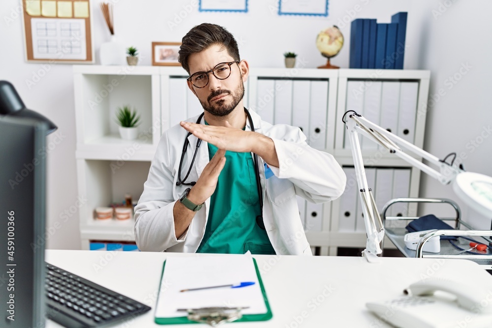 Young man with beard wearing doctor uniform and stethoscope at the clinic doing time out gesture with hands, frustrated and serious face