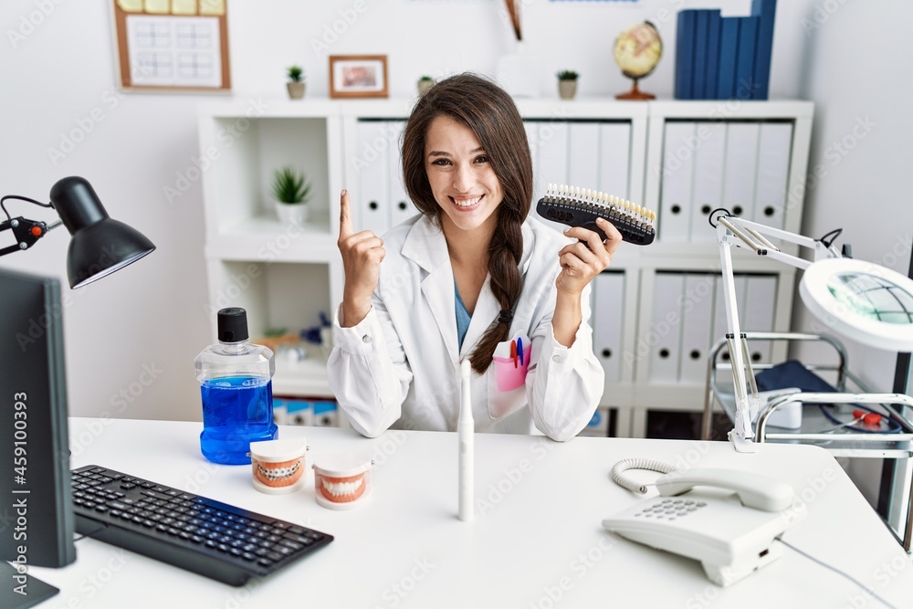 Young dentist woman comparing teeth whitening smiling with an idea or question pointing finger with happy face, number one Young dentist woman comparing teeth whitening smiling with an idea or question pointing finger with happy face, number one