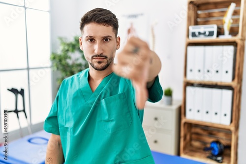 Young physiotherapist man working at pain recovery clinic looking unhappy and angry showing rejection and negative with thumbs down gesture. bad expression.