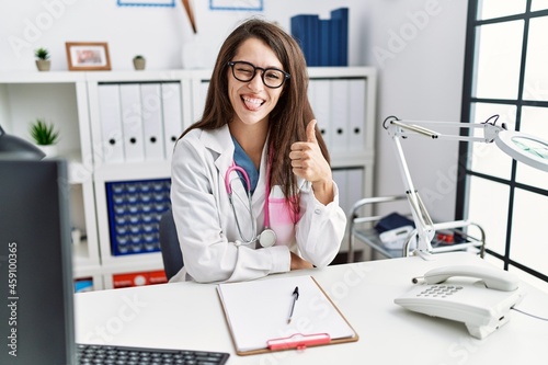 Young doctor woman wearing doctor uniform and stethoscope at the clinic doing happy thumbs up gesture with hand. approving expression looking at the camera showing success.