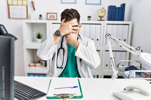 Young man with beard wearing doctor uniform and stethoscope at the clinic covering eyes and mouth with hands  surprised and shocked. hiding emotion