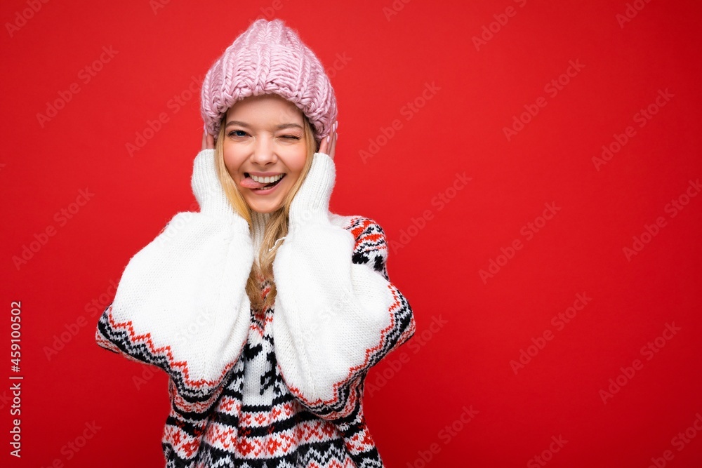 Photo of beautiful happy funny joyful young blonde woman isolated over red background wall wearing winter warm sweater and pink hat looking at camera winking and showing tongue Photo of beautiful happy funny joyful young blonde woman isolated over red background wall wearing winter warm sweater and pink hat looking at camera winking and showing tongue