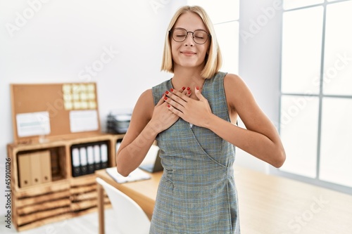 Young caucasian woman working at the office wearing glasses smiling with hands on chest with closed eyes and grateful gesture on face. health concept.