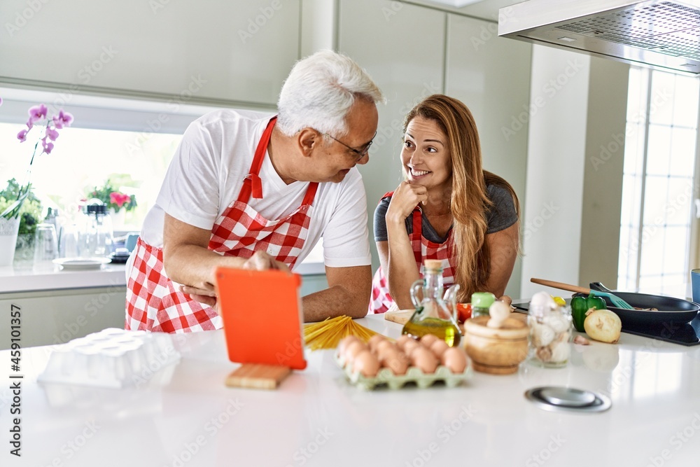 Middle age hispanic couple smiling happy cooking and using touchpad at the kitchen.