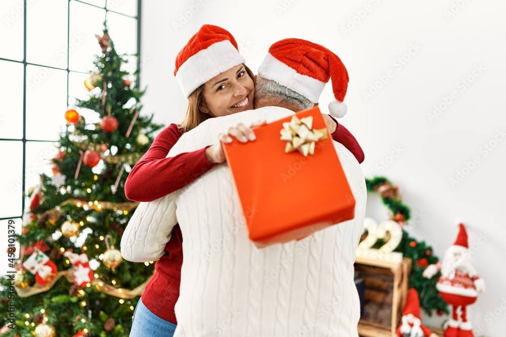 Middle age hispanic couple smiling happy and hugging holding christmas gift at home.