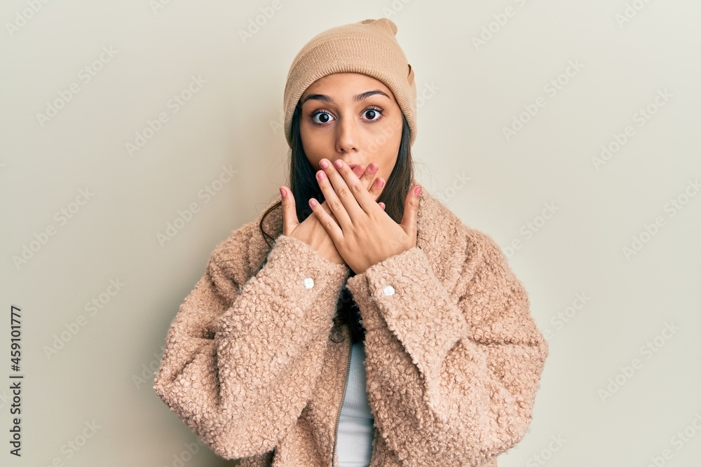 Young hispanic woman wearing wool sweater and winter hat shocked covering mouth with hands for mistake. secret concept.
