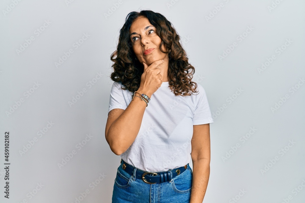 Middle age hispanic woman wearing casual white t shirt with hand on chin thinking about question, pensive expression. smiling with thoughtful face. doubt concept. Middle age hispanic woman wearing casual white t shirt with hand on chin thinking about question, pensive expression. smiling with thoughtful face. doubt concept.