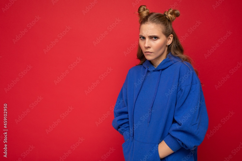 Portrait of touchy sad young beautiful winsome blonde woman with two horns with sincere emotions wearing stylish bright blue hoodie isolated on red background with copy space Portrait of touchy sad young beautiful winsome blonde woman with two horns with sincere emotions wearing stylish bright blue hoodie isolated on red background with copy space