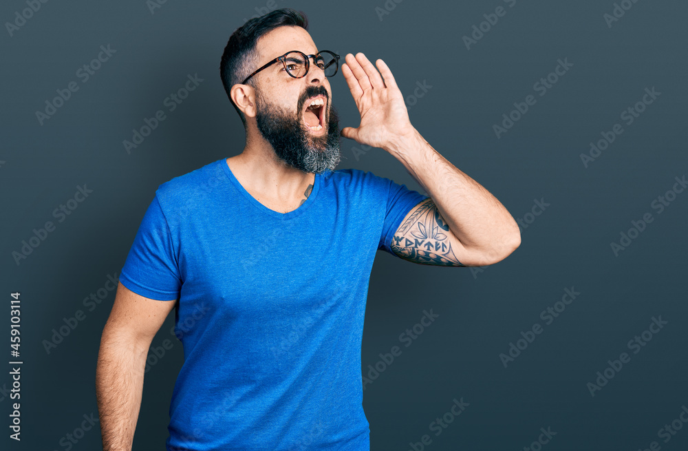 Hispanic man with beard wearing casual t shirt and glasses shouting and screaming loud to side with hand on mouth. communication concept. Hispanic man with beard wearing casual t shirt and glasses shouting and screaming loud to side with hand on mouth. communication concept.