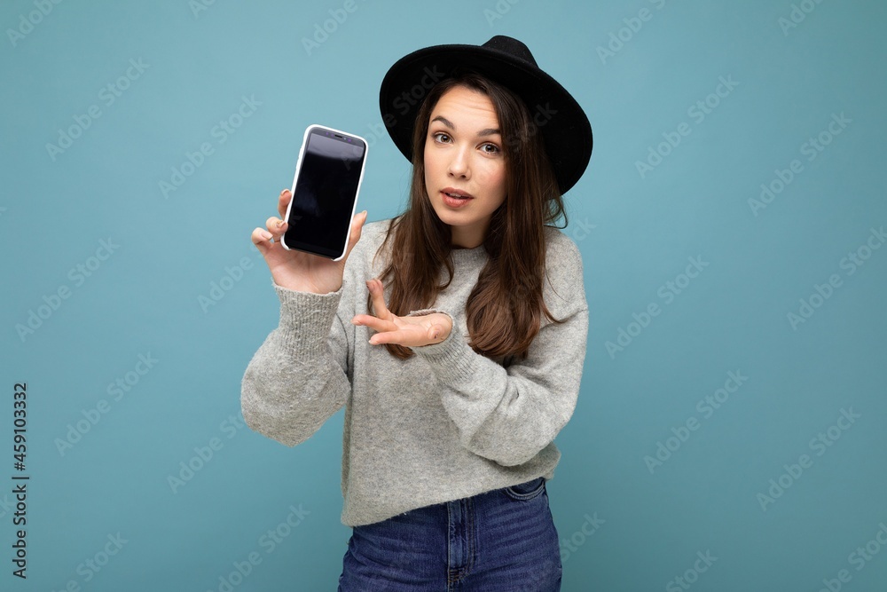 Beautiful young asking dissatisfied brunette woman wearing black hat and grey sweater holding smartphone looking at camera isolated on background Beautiful young asking dissatisfied brunette woman wearing black hat and grey sweater holding smartphone looking at camera isolated on background
