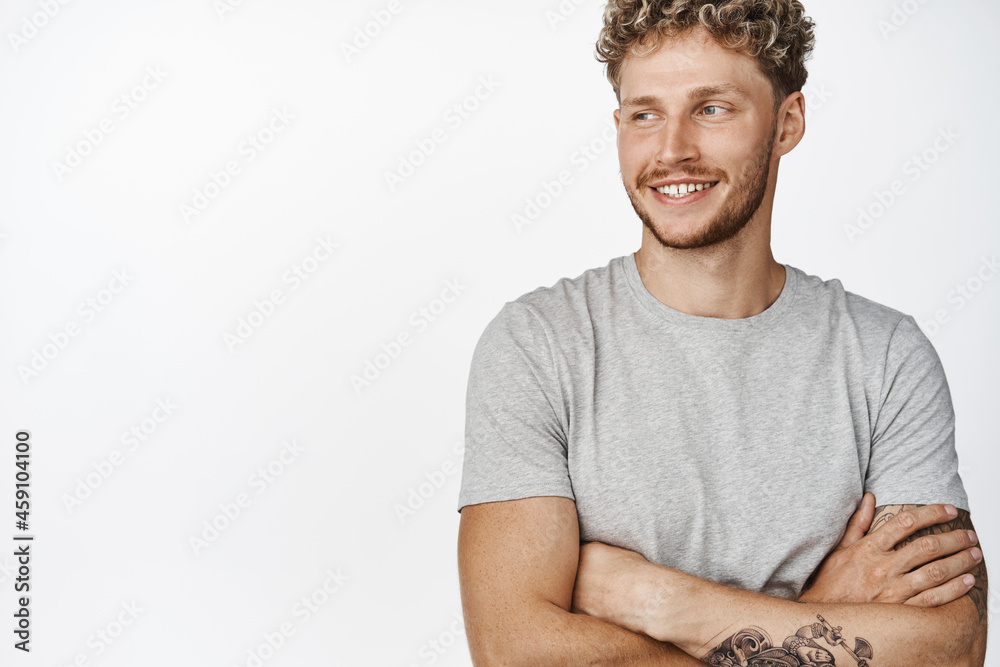 Close up shot of handsome young blond man with tattoos, bristle and happy smile, cross arms on chest, standing in relaxed pose, wearing grey t-shirt, white background