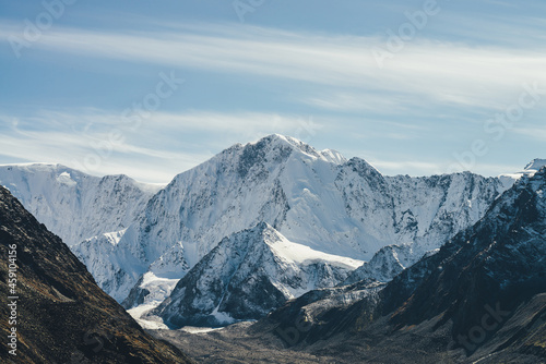 Alpine landscape with high snowy mountain with peaked top under cirrus clouds in sky. Atmospheric view to big snow covered mountains in sunshine. Black rocks and white-snow pointy peak in sunlight.