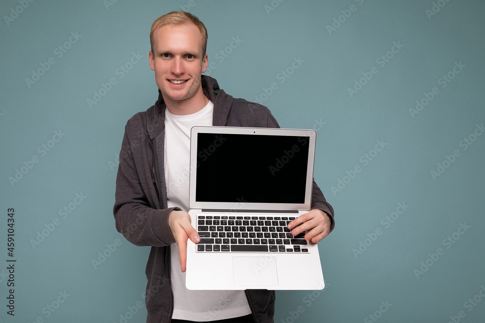 Photo of handsome blond man holding computer laptop looking at camera isolated over blue wall background wearing white t-shirt and grey sweater Photo of handsome blond man holding computer laptop looking at camera isolated over blue wall background wearing white t-shirt and grey sweater