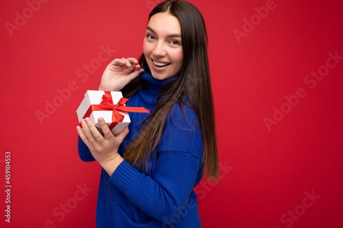 Shot of beautiful positive smiling young brunette woman isolated over red background wall wearing blue casual sweater holding white gift box with red ribbon and looking at camera