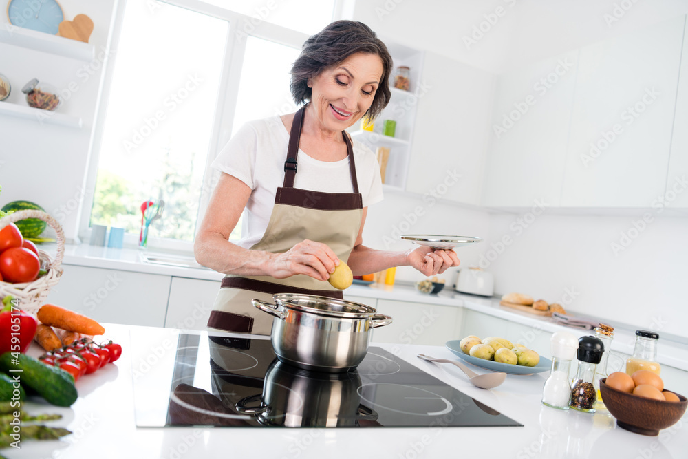 Photo of cheerful happy positive joyful old woman cook food potato smile indoors inside house home kitchen Photo of cheerful happy positive joyful old woman cook food potato smile indoors inside house home kitchen