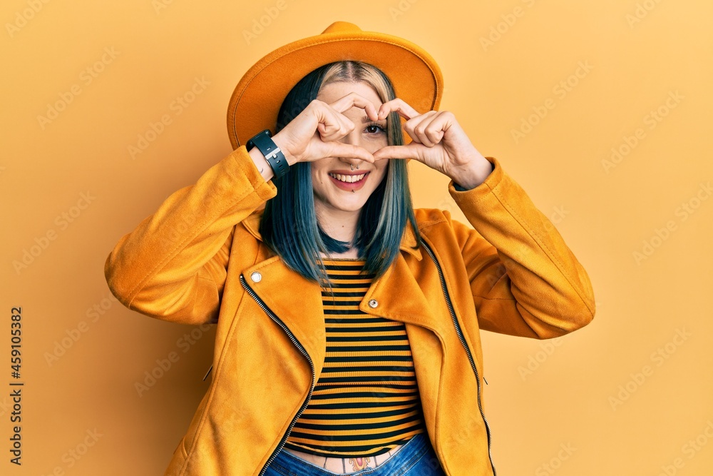 Young modern girl wearing yellow hat and leather jacket doing heart shape with hand and fingers smiling looking through sign