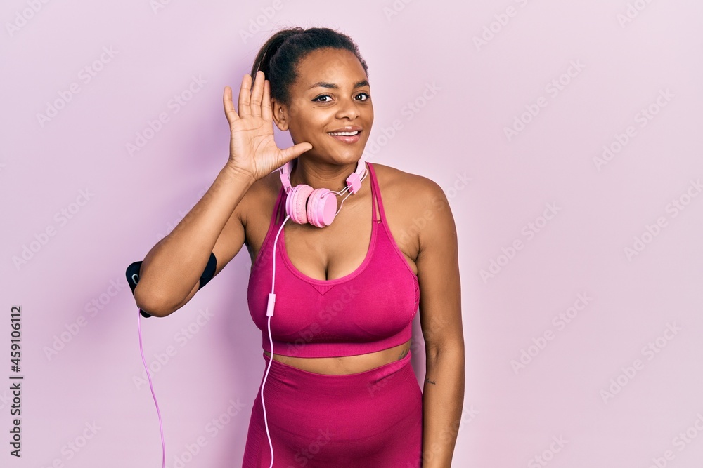 Young african american girl wearing gym clothes and using headphones smiling with hand over ear listening an hearing to rumor or gossip. deafness concept.