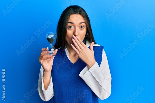 Young hispanic girl holding sand clock covering mouth with hand, shocked and afraid for mistake. surprised expression