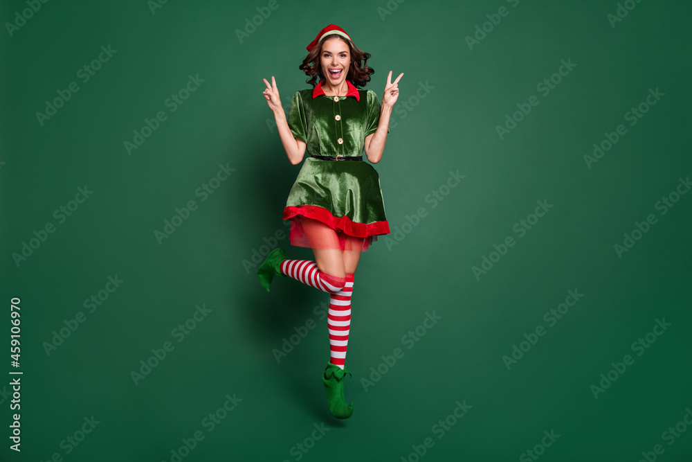 Full length body size view of pretty trendy cheerful girl jumping showing v-sign isolated over green color background