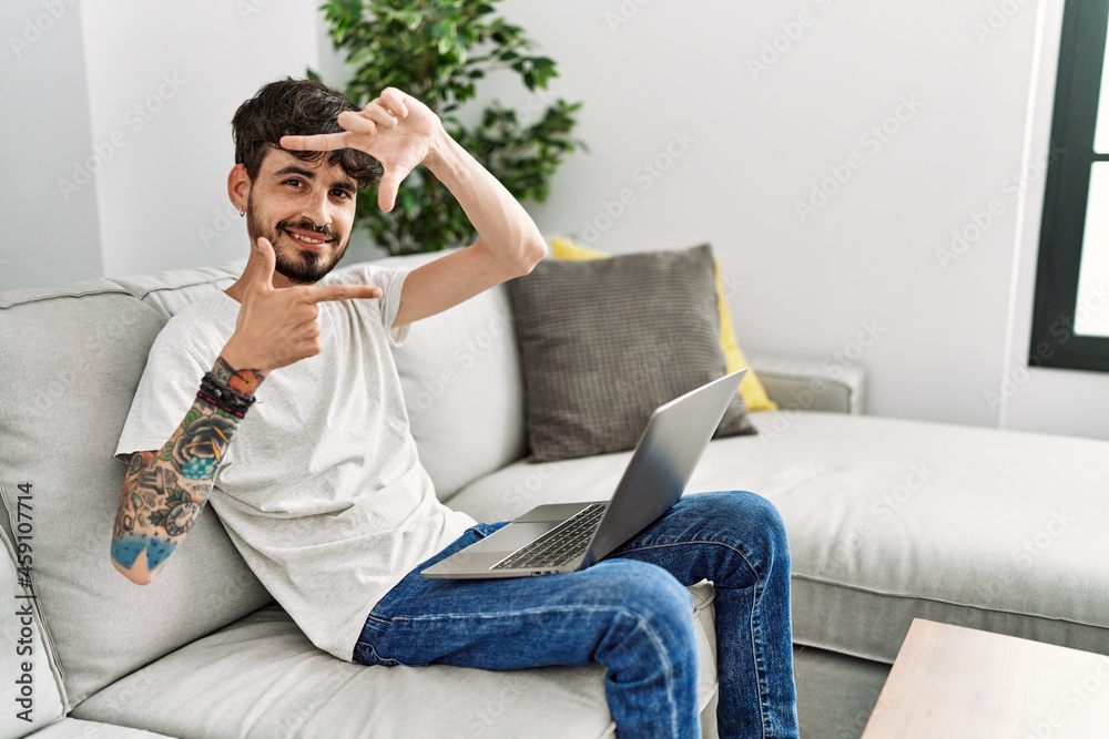 Fototapeta premium Hispanic man with beard sitting on the sofa smiling making frame with hands and fingers with happy face. creativity and photography concept.