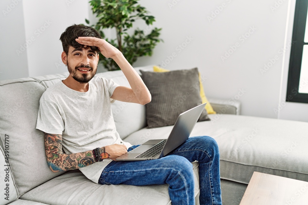 Hispanic man with beard sitting on the sofa very happy and smiling looking far away with hand over head. searching concept.