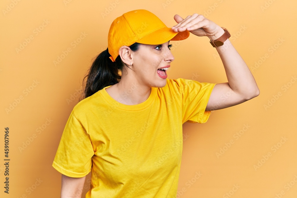 Young hispanic woman wearing delivery uniform and cap very happy and smiling looking far away with hand over head. searching concept.