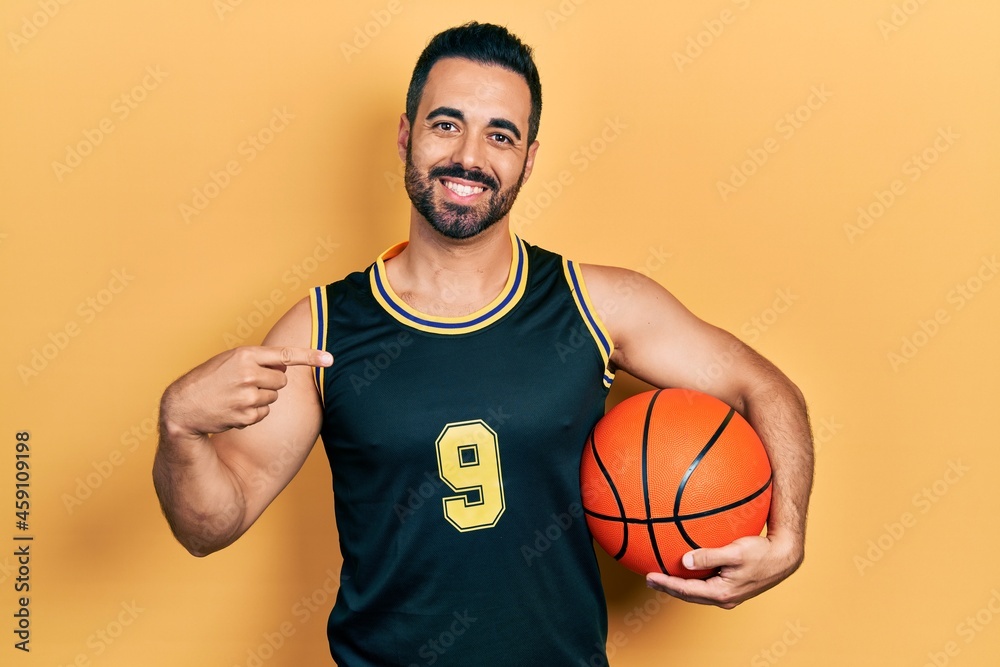 Handsome hispanic man with beard holding basketball ball pointing finger to one self smiling happy and proud