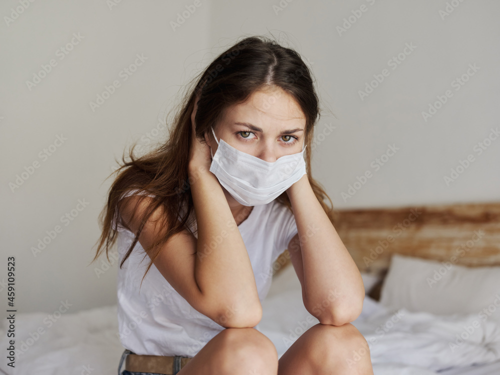 red-haired woman in medical mask sitting on bed in isolated room