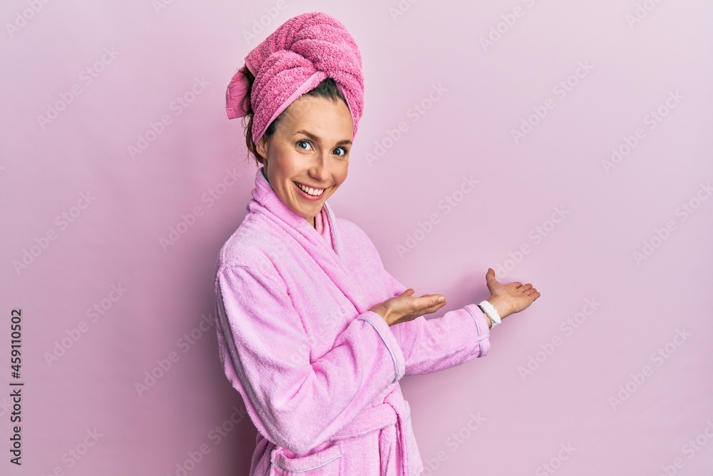 Young blonde woman wearing shower towel cap and bathrobe inviting to enter smiling natural with open hand Young blonde woman wearing shower towel cap and bathrobe inviting to enter smiling natural with open hand