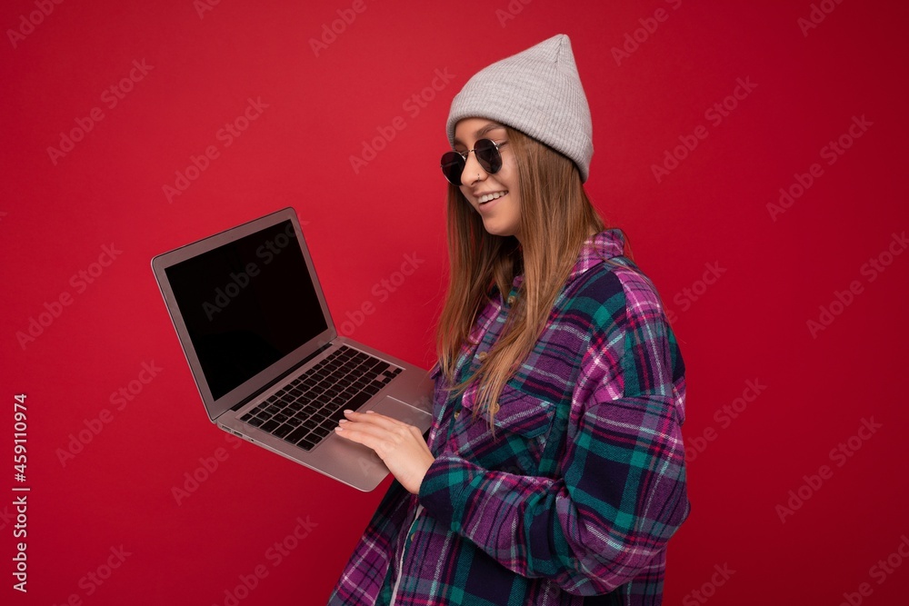 Side profile photo of beautiful young woman holding computer wearing casual smart clothes sunglasses and hat looking at monitor screen isolated on red background Side profile photo of beautiful young woman holding computer wearing casual smart clothes sunglasses and hat looking at monitor screen isolated on red background