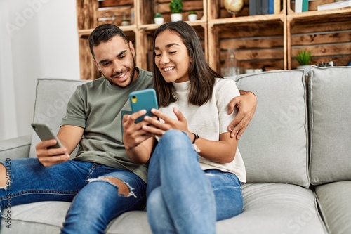 Young latin couple using smartphone sitting on the sofa at home.