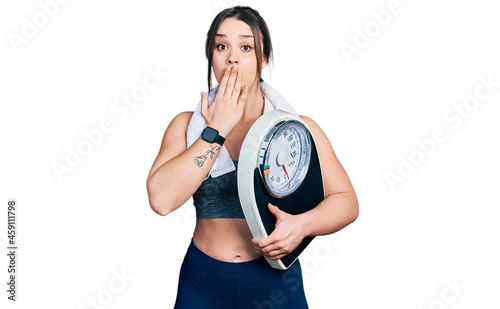 Young hispanic girl wearing sportswear holding weighing machine covering mouth with hand  shocked and afraid for mistake. surprised expression