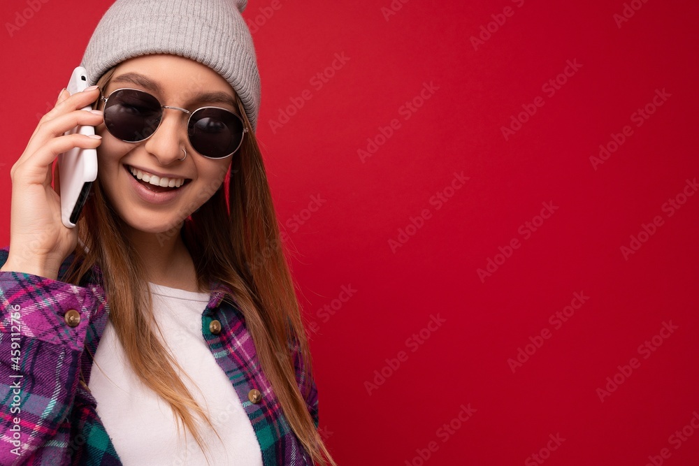 Closeup photo of beautiful happy positive young blonde woman wearing hipster purple shirt and casual white t-shirt grey hat and sunglasses isolated over red background holding in hand and talking on