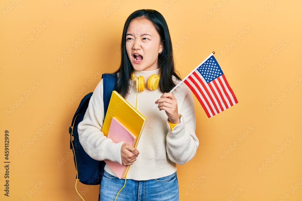 Young chinese girl exchange student holding america flag angry and mad screaming frustrated and furious, shouting with anger. rage and aggressive concept.