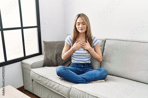 Asian young woman sitting on the sofa at home smiling with hands on chest with closed eyes and grateful gesture on face. health concept.