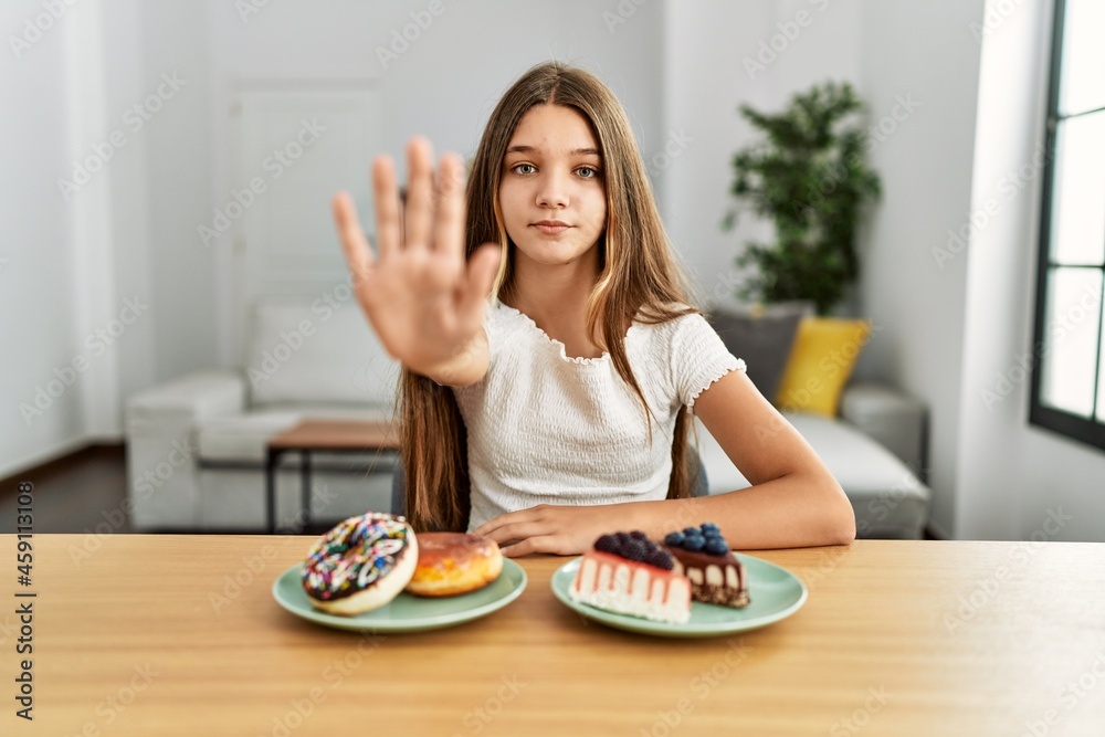 Young brunette teenager eating cakes and sweets with open hand doing stop sign with serious and confident expression, defense gesture Young brunette teenager eating cakes and sweets with open hand doing stop sign with serious and confident expression, defense gesture