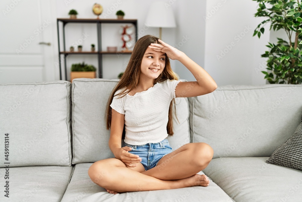 Young brunette teenager sitting on the sofa at home very happy and smiling looking far away with hand over head. searching concept.