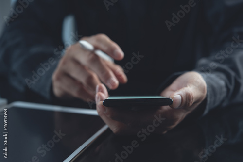 Close up of man hand holding and using mobile phone with digital tablet on table at office, business background, dark tone
