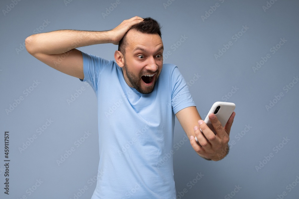 Furious shocked handsome young bearded brunet man wearing everyday blue t-shirt isolated over blue background holding and using mobile phone communication online on the internet looking at gadjet Furious shocked handsome young bearded brunet man wearing everyday blue t-shirt isolated over blue background holding and using mobile phone communication online on the internet looking at gadjet