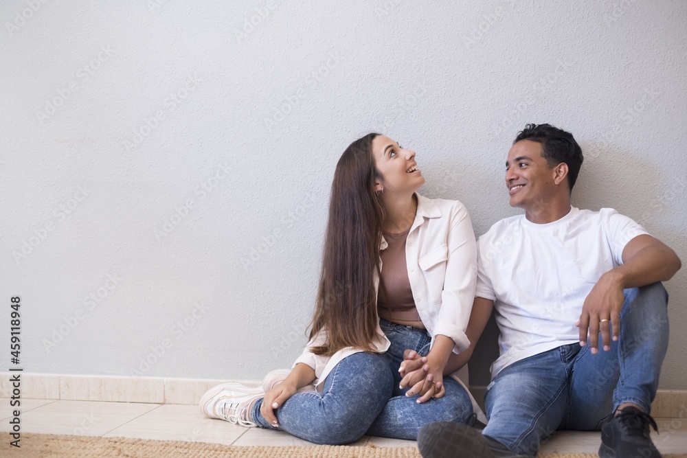 Young couple sitting on floor of living room holding hands and leaning on wall. Couple spending quality time at home. Cheerful couple sitting on floor of new house Young couple sitting on floor of living room holding hands and leaning on wall. Couple spending quality time at home. Cheerful couple sitting on floor of new house
