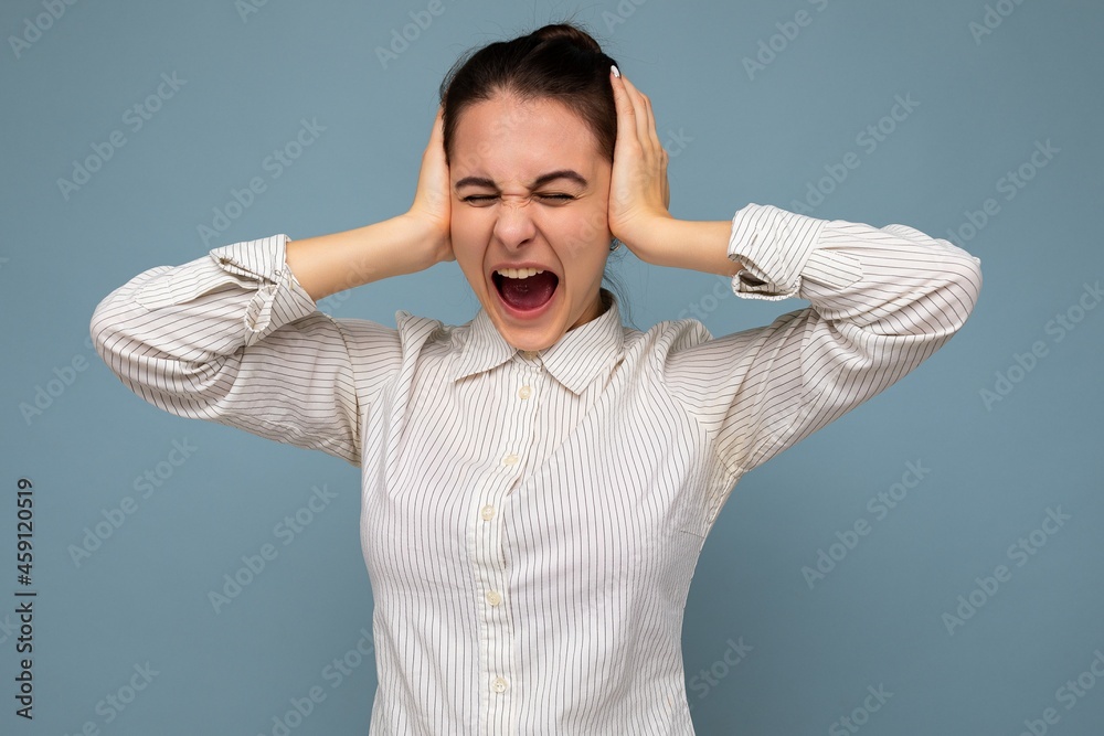 Portrait of emotional young beautiful brunette woman with sincere emotions wearing white shirt isolated over blue background with copy space and covering ears with hands and shouting
