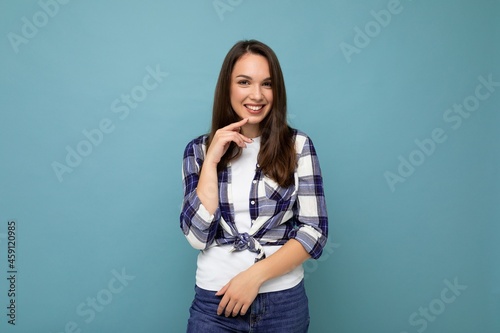 Young positive happy smiling beautiful winsom brunette woman with sincere emotions wearing check shirt poising isolated over blue background with copy space