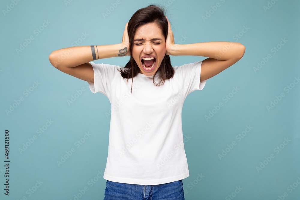 Portrait of emotional young beautiful brunette woman with sincere emotions wearing casual white t-shirt for mockup isolated on blue background with empty space and covering ears with hands and Portrait of emotional young beautiful brunette woman with sincere emotions wearing casual white t-shirt for mockup isolated on blue background with empty space and covering ears with hands and