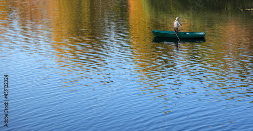 Fisherman with spinning stands in wooden boat against background of reflection of autumn foliage in blue water. Fishing in autumn on river.