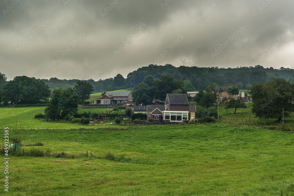 Houses in a hilly landscape with trees and meadows under a cloudy sky.