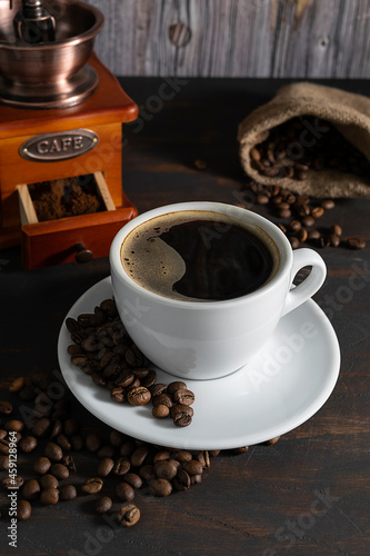 A white cup with black coffee  coffee beans on a saucer. A coffee grinder and a bag of coffee beans in the background