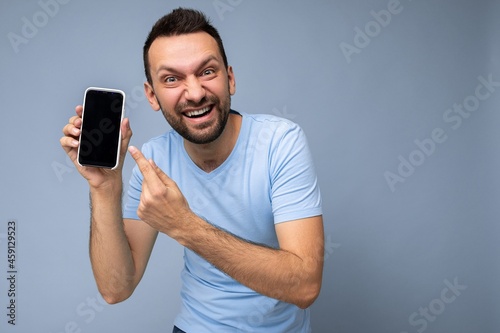 Overemotional handsome young unshaven brunet man wearing everyday blue t-shirt isolated over blue background holding and showing mobile phone with empty display for cutout looking at camera and